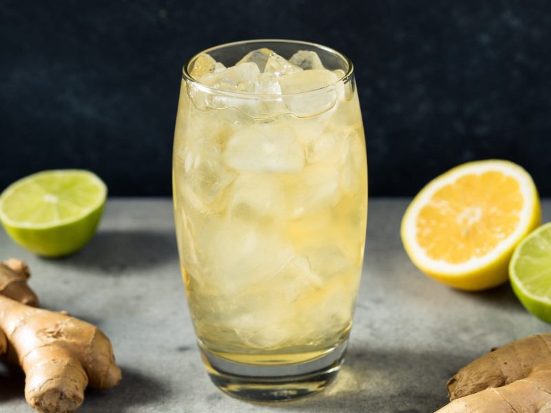 Ein Glas mit Ginger Beer und Eis auf einem grauen Tisch mit Ingwer und Zitrusfrüchten.