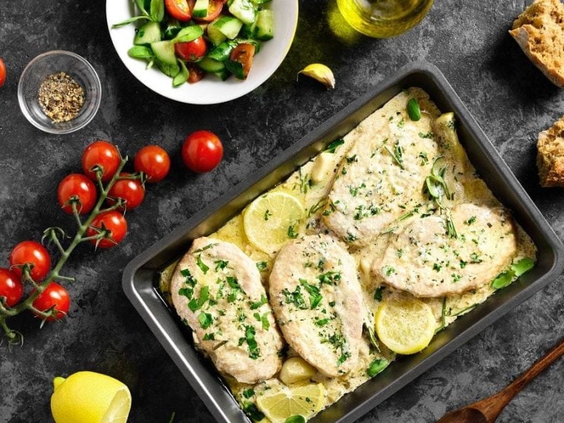 Eine Auflaufform mit Hähnchen in zitroniger Knoblauchsahne steht auf einem Tisch. Drumherum liegen Tomaten und andere Lebensmittel. Sowie ein frischer grüner Salat.