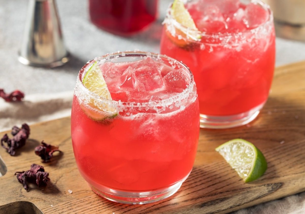 Zwei Gläser mit dem Hibiskus-Mezcal-Margarita auf einem Holzbrett mit Hibiskus-Blüten und Limetten.