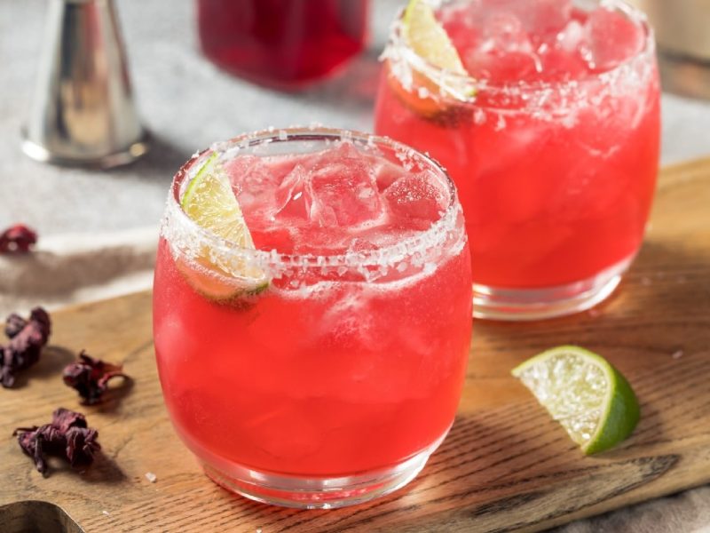 Zwei Gläser mit dem Hibiskus-Mezcal-Margarita auf einem Holzbrett mit Hibiskus-Blüten und Limetten.