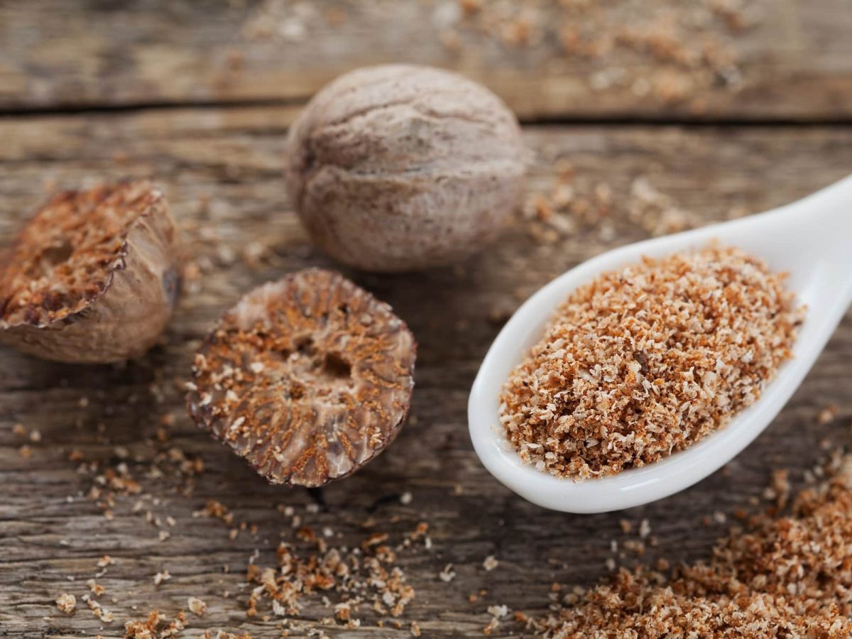 Auf einem Untergrund aus Holz liegen ein ganze Muskatnuss, zwei halbe Muskatnüsse sowie in Löffel mit geriebenem Muskat.