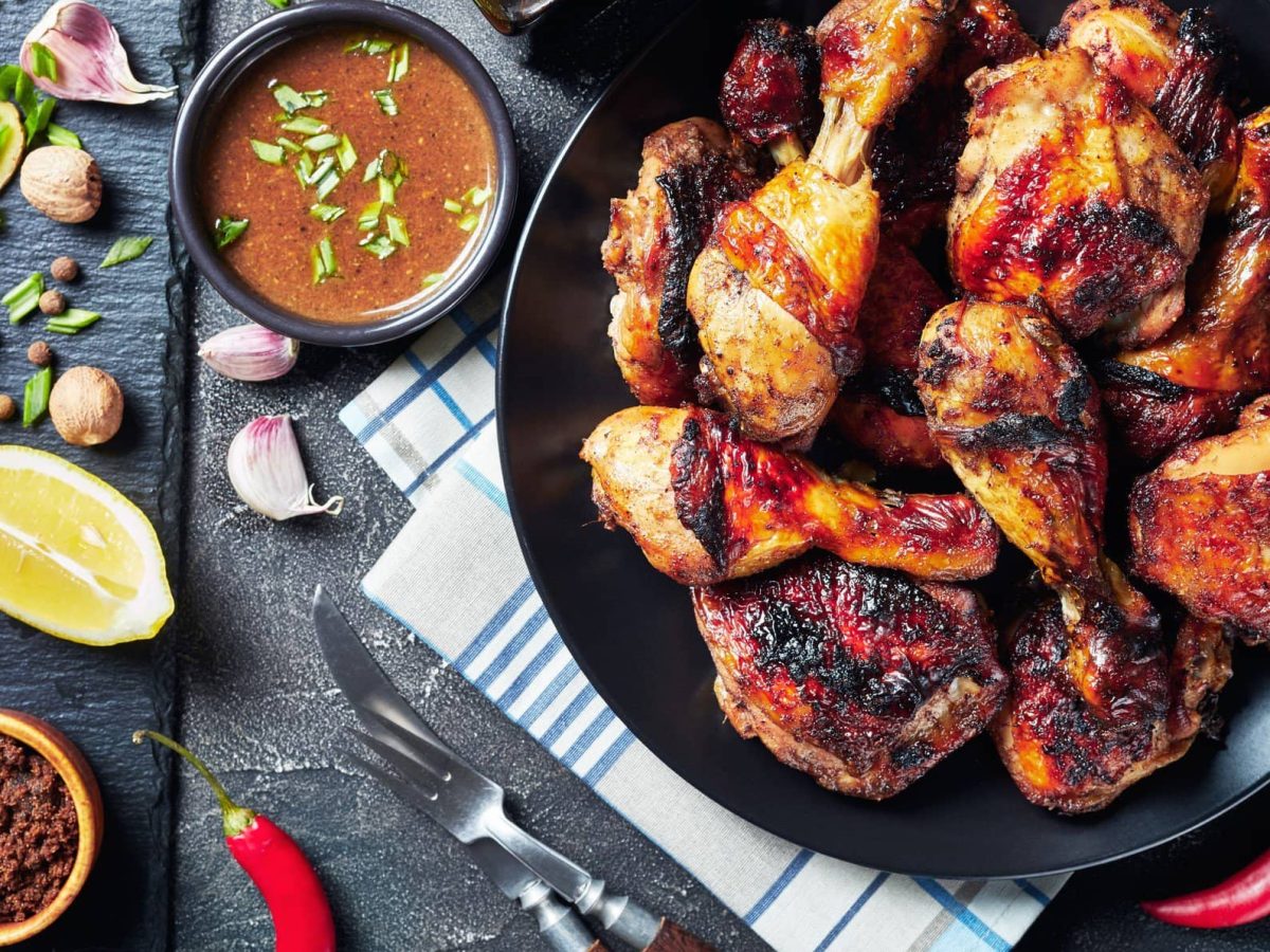 Jerk Chicken auf schwarzem Teller auf Tuch auf grauem Untergrund. Daneben Gewürz, Chilis, Limettenspalten, Knoblauch und Lauchstücken. Draufsicht.und