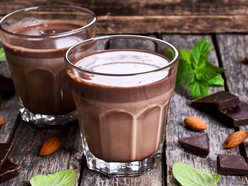 Zweimal Kakao-Shake mit Johannisbeeren und Apfelsaft in Glasern auf grauem Tisch vor Holz. Im Bild Minze, Schokolade und Mandeln. Frontalansicht.