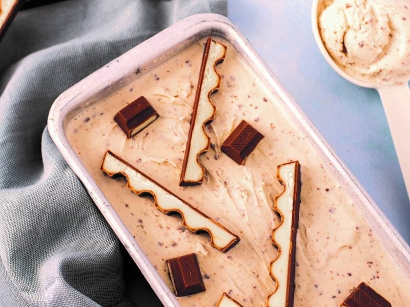 Kinderschokolade-Eis in einer Kastenform, mit Stückchen von Kinderschokolade belegt, daneben ein Eislöffel.