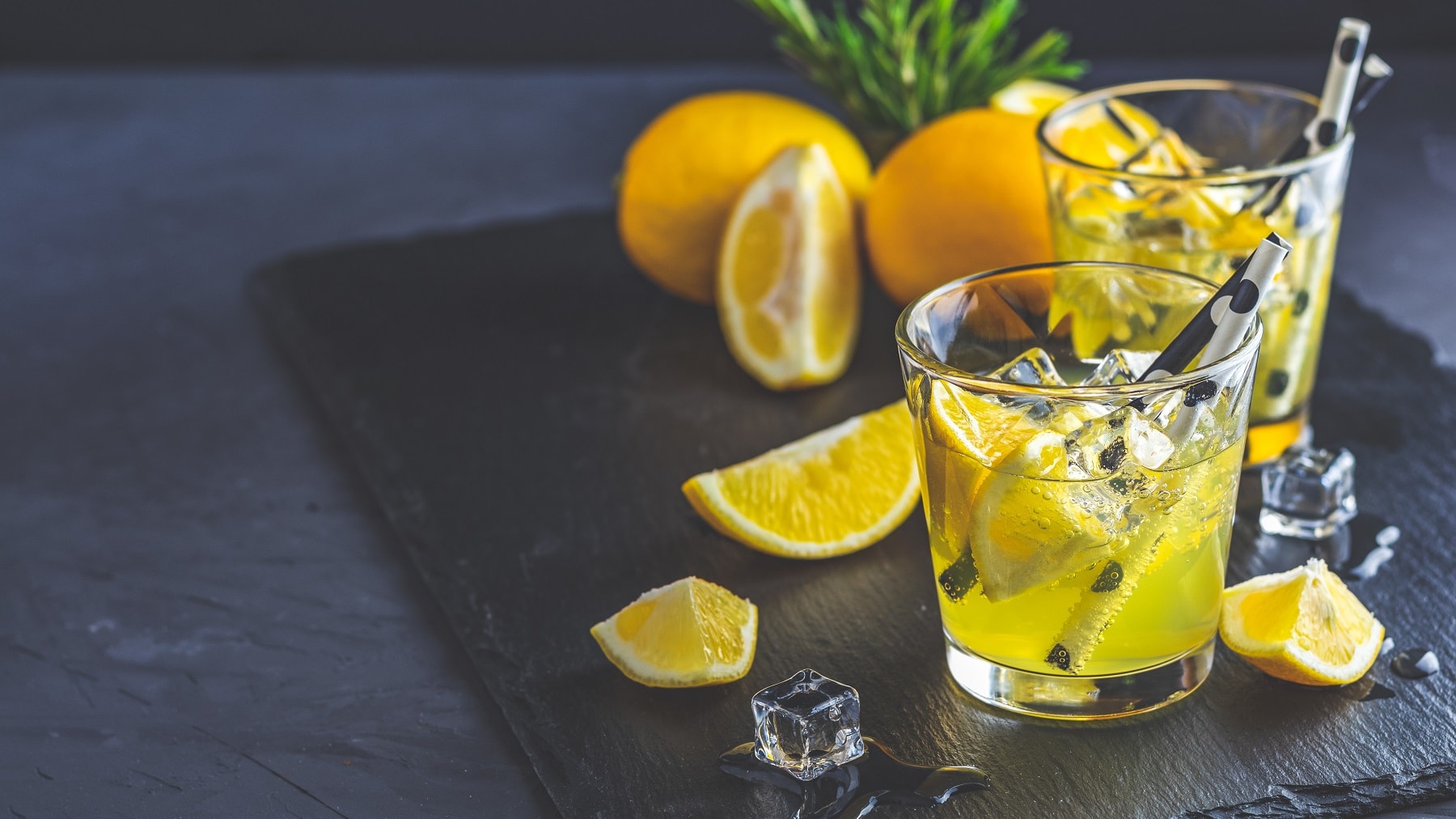 zwei Gläser Limoncello Tonic mit Eiswürfeln und Zironenstücken, darin Strohhalme. Drumherum frische angeschnittene Zitronen.