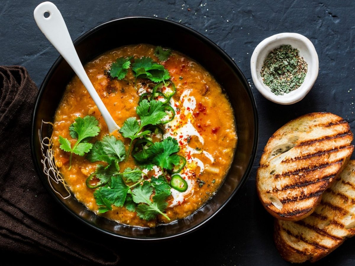 Eine schwarze Schüssel Linsen-Kokos-Suppe mit einem weißen Keramiklöffel darin in der Draufsicht, daneben eine Schale Gewürze, zwei getoastete Brotscheiben und ein braunes Geschirrtuch.