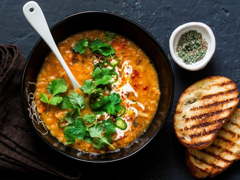 Eine schwarze Schüssel Linsen-Kokos-Suppe mit einem weißen Keramiklöffel darin in der Draufsicht, daneben eine Schale Gewürze, zwei getoastete Brotscheiben und ein braunes Geschirrtuch.