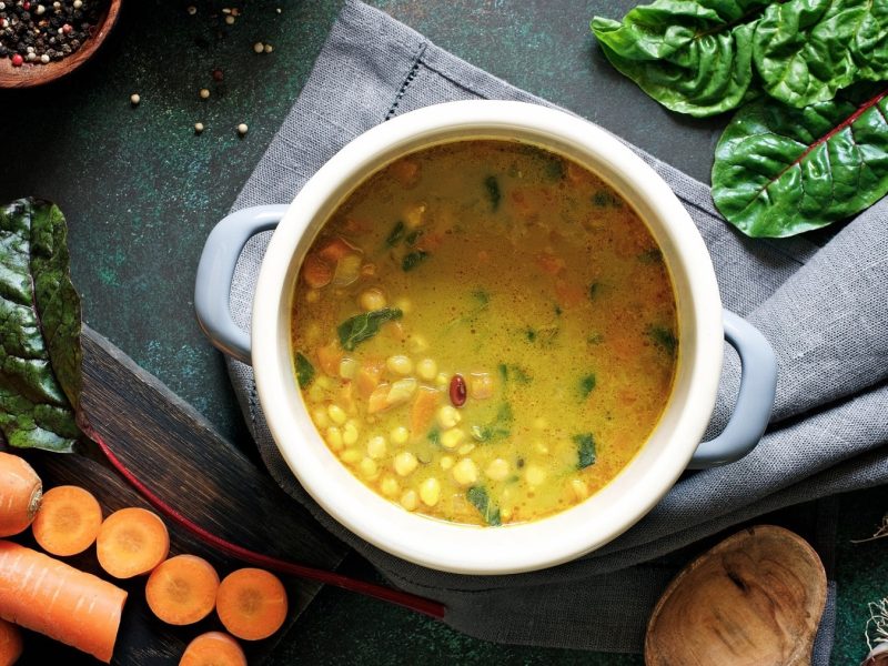 Eine Suppenterrine mit Linsensuppe mit Mangold und Kichererbsen in der Draufsicht, drumherum liegen frischer Mangold und Möhren sowie Knoblauch.