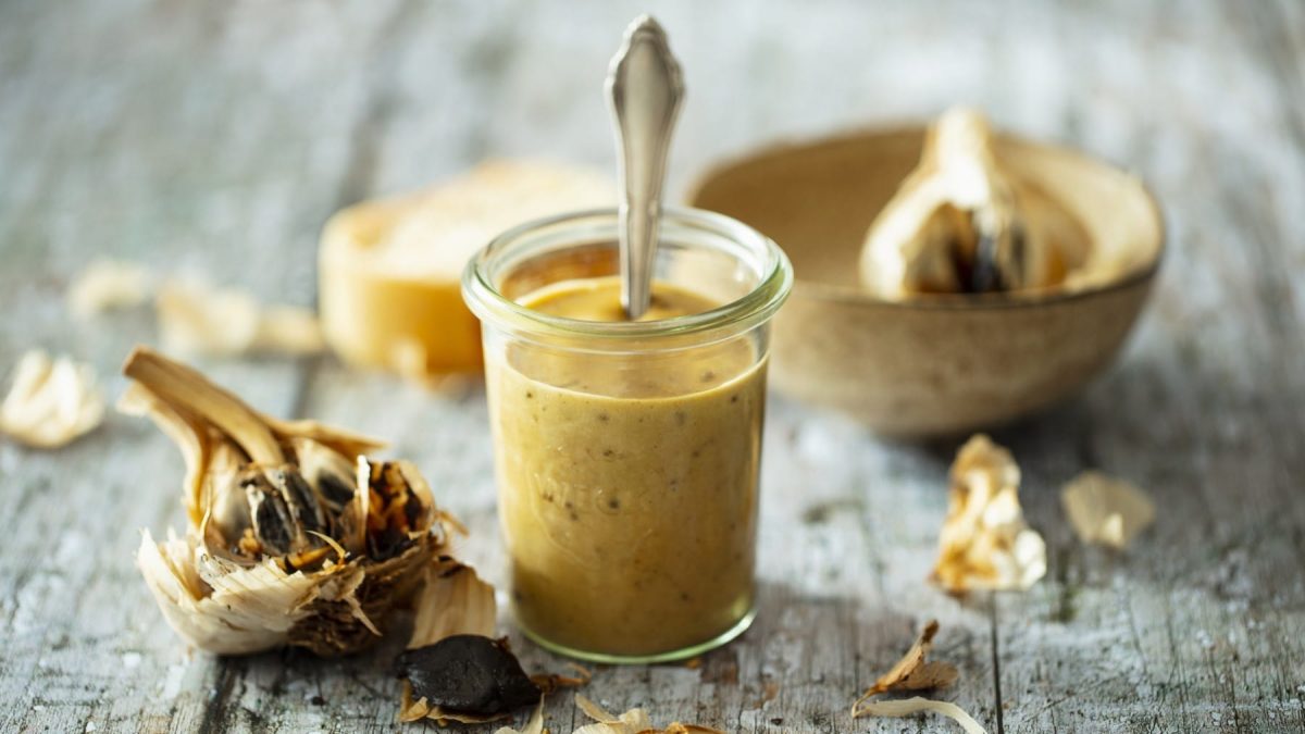 Mayonnaise aus fermentiertem Knoblauch, serviert in einem kleinen Einweckglas mit einem feinen Löffel, und präsentiert umgeben von angeröstetem Knoblauch auf einem hell-grauen Tisch.