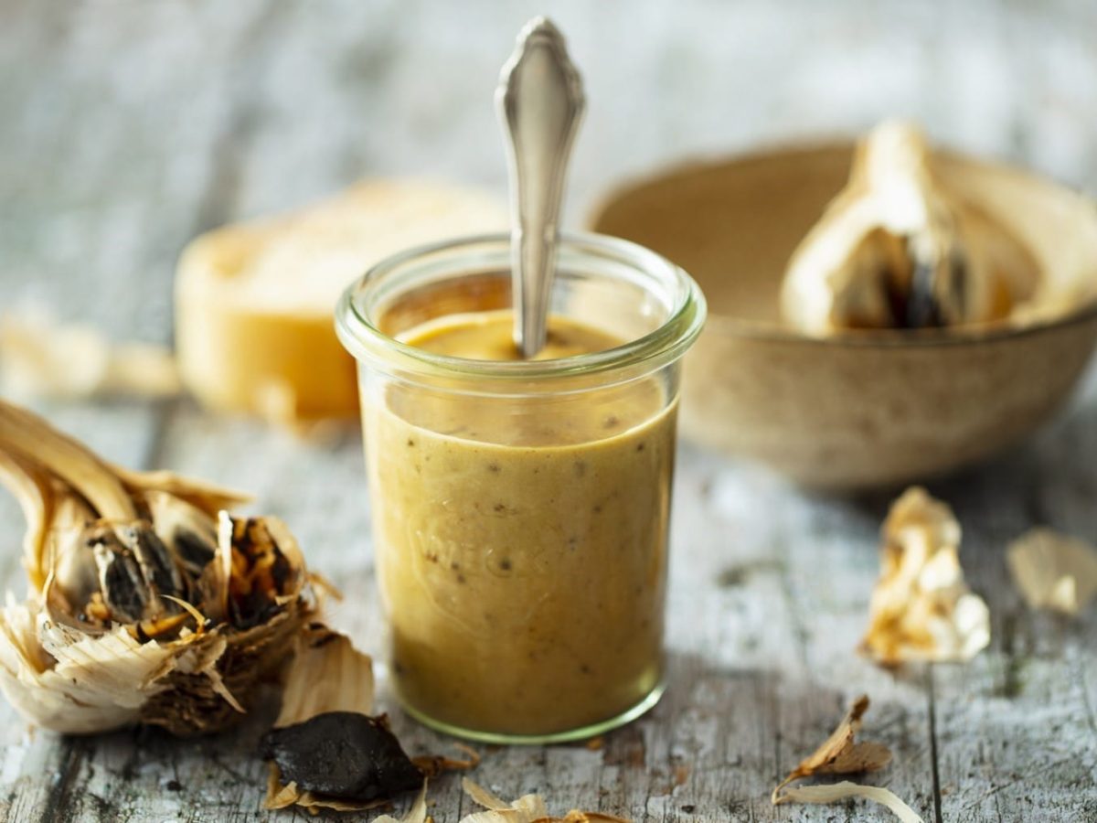 Mayonnaise aus fermentiertem Knoblauch, serviert in einem kleinen Einweckglas mit einem feinen Löffel, und präsentiert umgeben von angeröstetem Knoblauch auf einem hell-grauen Tisch.