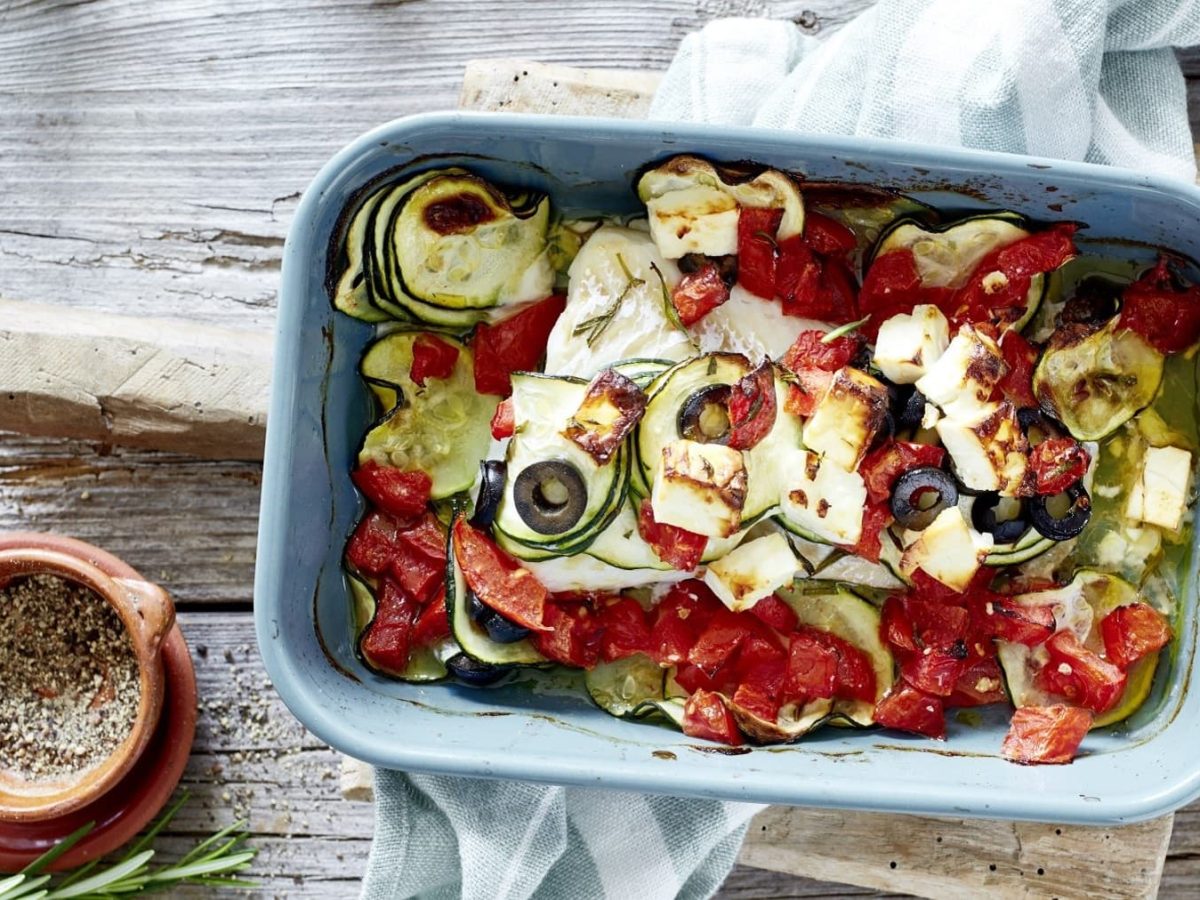 Bunter mediterraner Fischauflauf, bestehend aus leicht geschmorter Zucchini, schwarzen Oliven, Tomaten, gebackenem Feta und saftigem Kabeljaufilet, serviert in einer großen Form auf hölzernem Tisch.