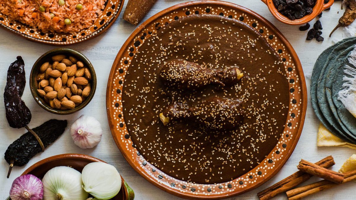 Mole poblano mit Hähnchen in Tonschale auf hellem Tisch mit Zutaten und Reis in Schalen und auf dem Tisch verteilt. Aufnahme aus der Vogelperspektive.