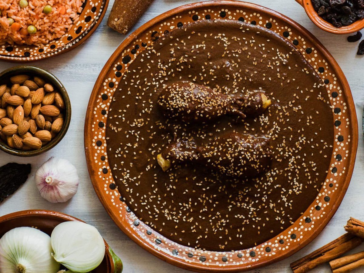 Mole poblano mit Hähnchen in Tonschale auf hellem Tisch mit Zutaten und Reis in Schalen und auf dem Tisch verteilt. Aufnahme aus der Vogelperspektive.