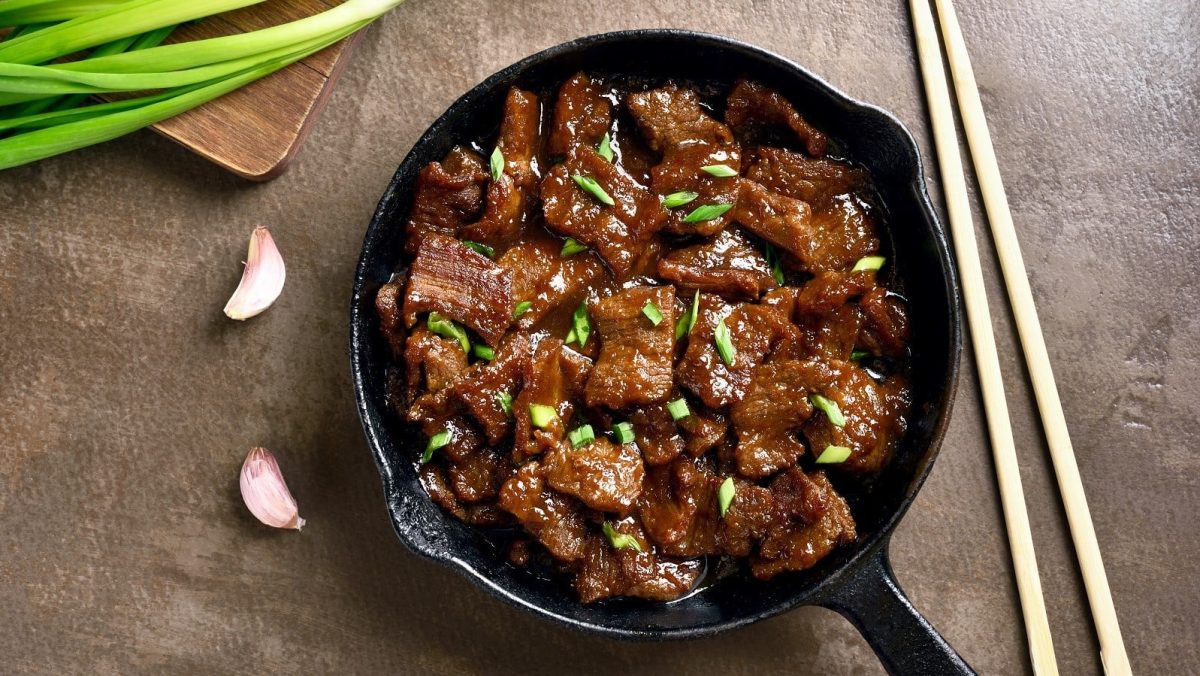 Eine Pfanne mit mongolischem Rindfleisch neben frischen Lauchzwiebeln und einem Paar Stäbchen