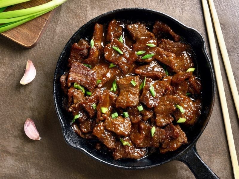 Eine Pfanne mit mongolischem Rindfleisch neben frischen Lauchzwiebeln und einem Paar Stäbchen