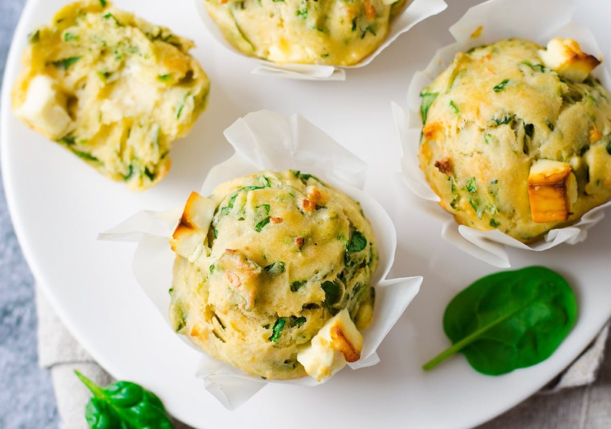 Ein weißer Teller auf dem die herzhaften Muffins mit Spinat und Feta liegen. Im Hintergrund ist eine grau marmorierte Wand und etwas Blattspinat zu erkennen.