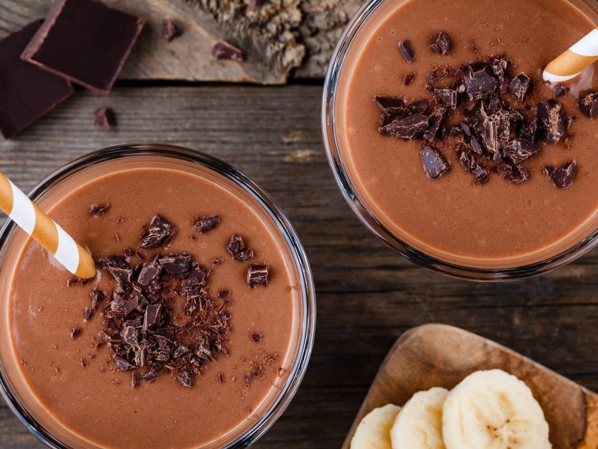 Pistazienmus-Smoothie mit Kakao, Bananen und Beeren in Gläsern mit Strohhalm auf dunklem Holz. Daneben Holzbrett mit Bananenscheiben. Oben Schokolade. Draufsicht.