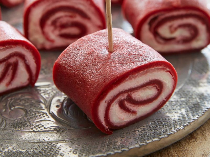 Mehrere Rote Bete-Creperöllchen auf kleinen Spießen auf einem dunklen Teller vor Holzhintergrund.