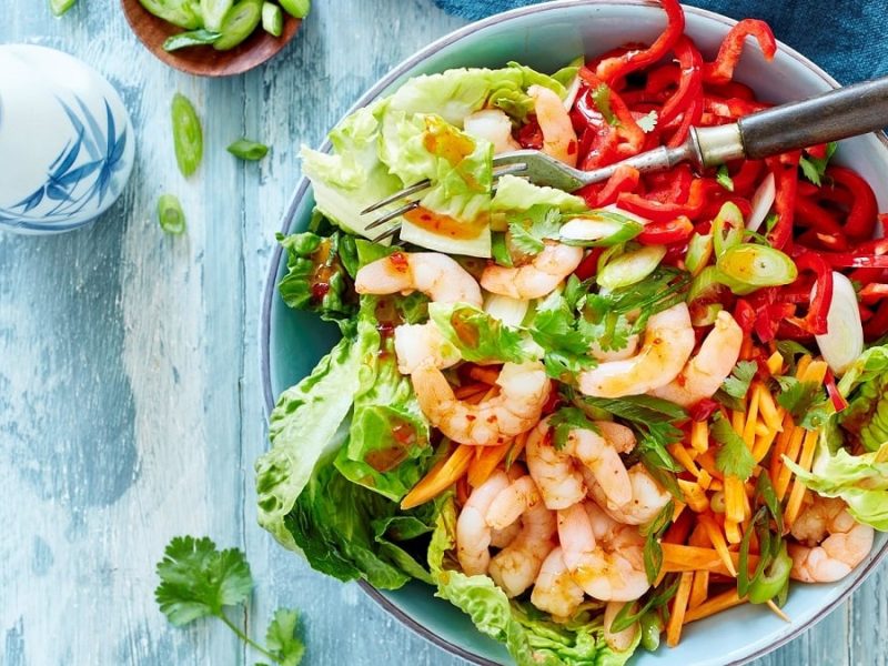 eine Schüssel Salat mit Shrimps und asiatischem Dressing steht auf einem hellblauen Untergrund aus Holz.