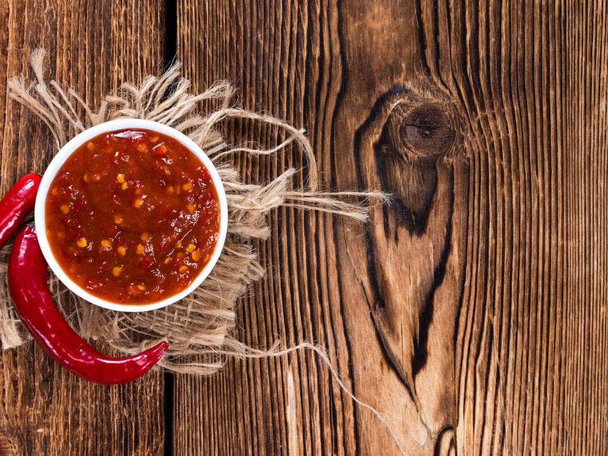 Selbst gemachtes Sambal Oelek in weißer Schale auf beigem Tuch auf Holztisch. Daneben Chilischoten. Aufnahme aus der Vogelperspektive.