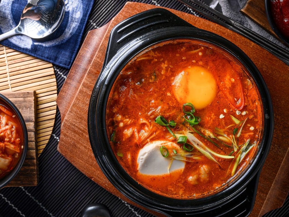 Ein schwarzer Topf mit Sundubu Jjigae neben Kimchi.