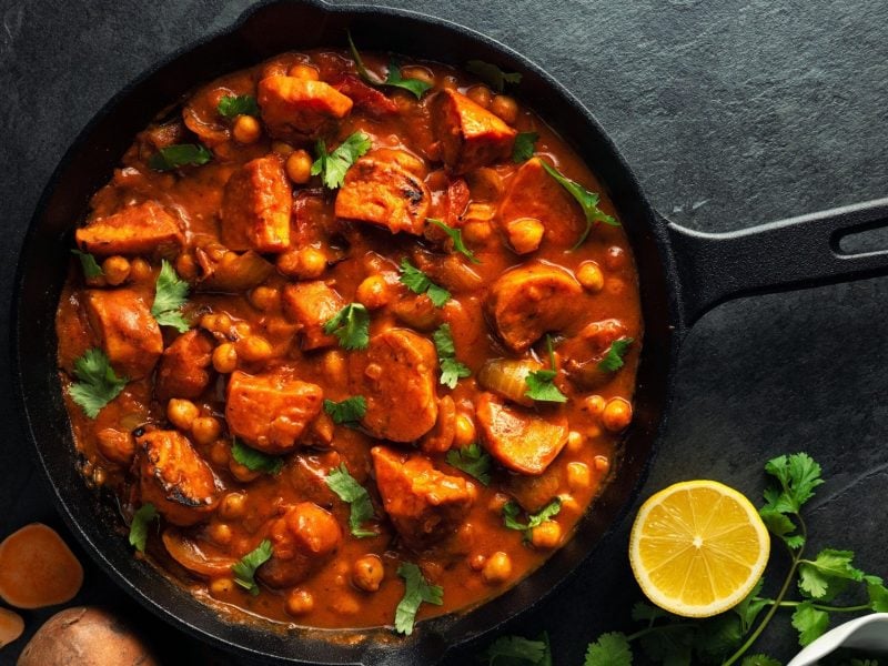 Eine Pfanne Tikka Masala mit Süßkartoffeln und Kichererbsen, bestreut mit frischem Koriander, in der Draufsicht, daneben eine halbe Zitrone.