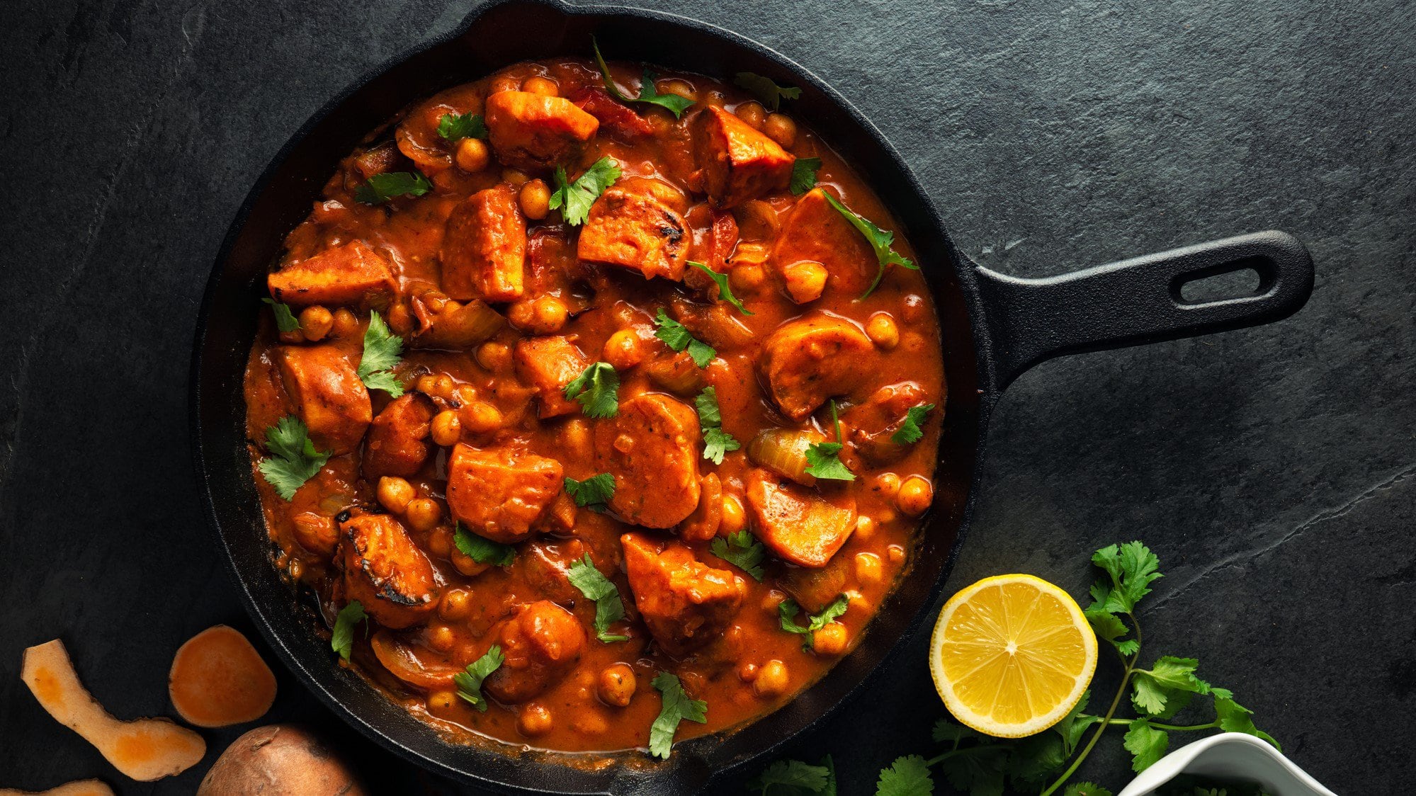 Eine Pfanne Tikka Masala mit Süßkartoffeln und Kichererbsen, bestreut mit frischem Koriander, in der Draufsicht, daneben eine halbe Zitrone.