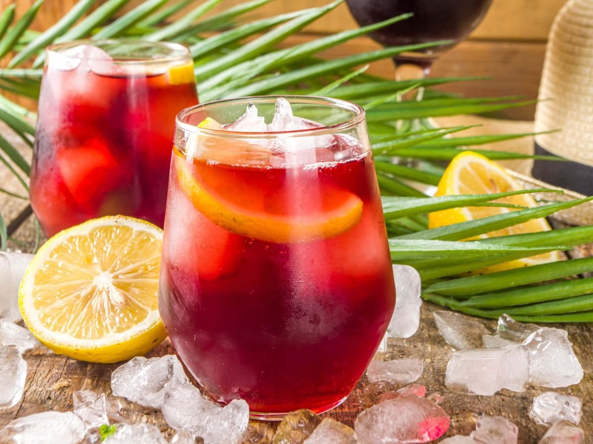 Tinto de Verano in zwei Gläsern auf Holztisch mit Eiswürfeln, Pflanzen, Zitronen und Strohhut. Im Hintergrund Rotweinglas erkennbar. Frontalansicht.