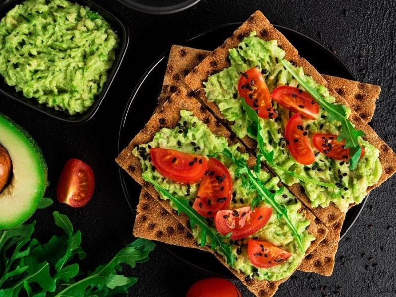 Zwei Tomaten-Knäcke mit Avocado-Rucola-Pesto liegen auf einem Holzbrettchen. Daneben liegen eine halbe Avocado mit Kern sowie ein paar Tomaten.