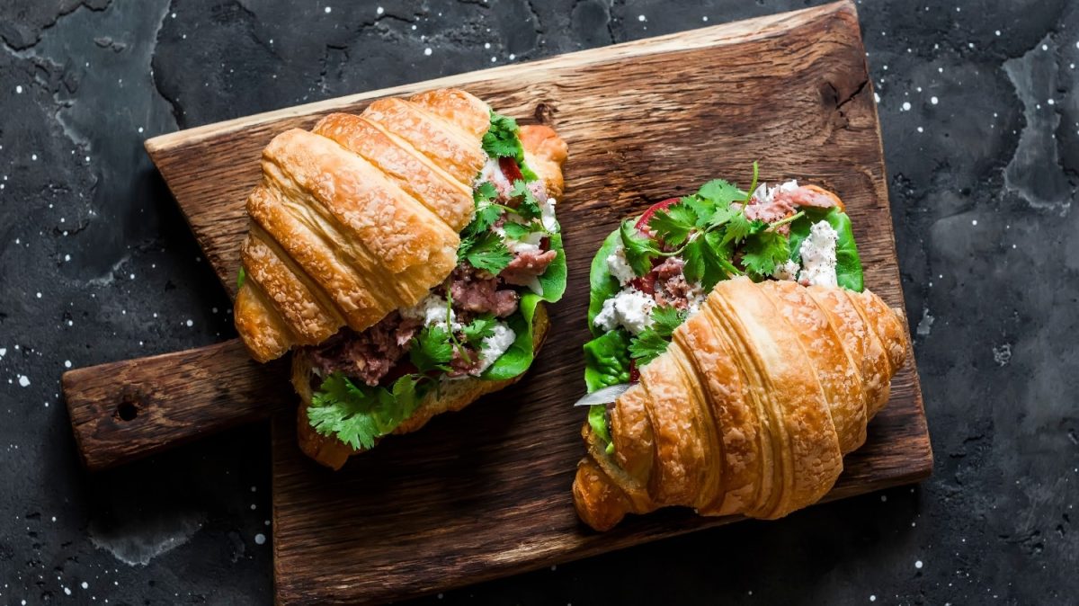 Zwei angeschnittene Croissants auf einem dunklem Holzbrett mit Koriander und Thunfisch.