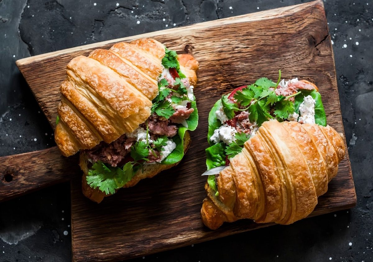 Zwei angeschnittene Croissants auf einem dunklem Holzbrett mit Koriander und Thunfisch.