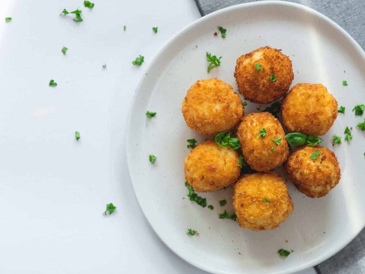 Ein Teller mit sieben veganen Arancini, bestreut mit frischer Petersilie und Basilikum. Draufsicht.