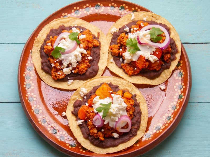 Drei vegane Hackfleisch-Tacos mit Bohnenmus, roten Zwiebeln, Koriander, saurer Sahne und Feta auf einem bunten Teller in der Draufsicht.