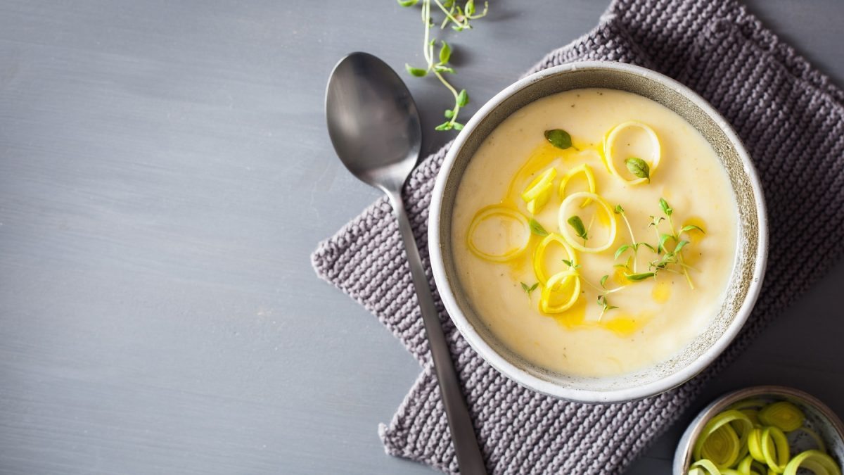 Eine Schüssel vegane Kartoffel-Lauch-Suppe mit frischem Lauch und Kräutern in der Draufsicht, daneben ein Löffel und ein grobmaschiges Geschirrtuch.