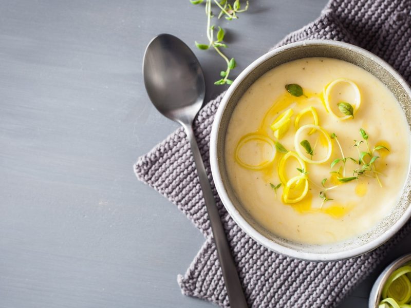 Eine Schüssel vegane Kartoffel-Lauch-Suppe mit frischem Lauch und Kräutern in der Draufsicht, daneben ein Löffel und ein grobmaschiges Geschirrtuch.