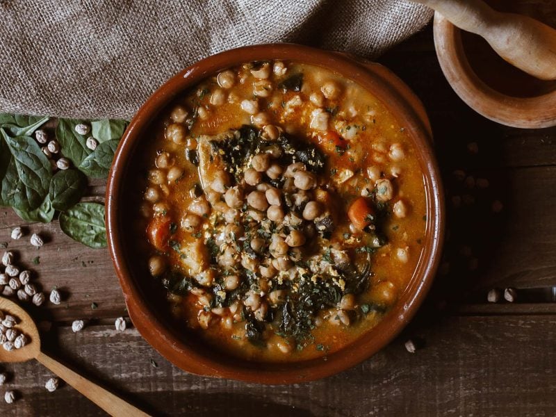 eine Tonschale mit veganem Mangold-Curry mit Kichererbsen in der Draufsicht, daneben Mörser und Stößel sowie ein Holzlöffel und Mangoldblätter.