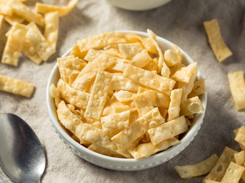 Wantan Chips aus selbst gemachtem Wantan Teig in weißer Schüssel auf Tuch. Im Bild Chips verteilt. Links Metalllöffel. Oben Schale angedeutet. Aufnahme aus der Vogelperspektive.