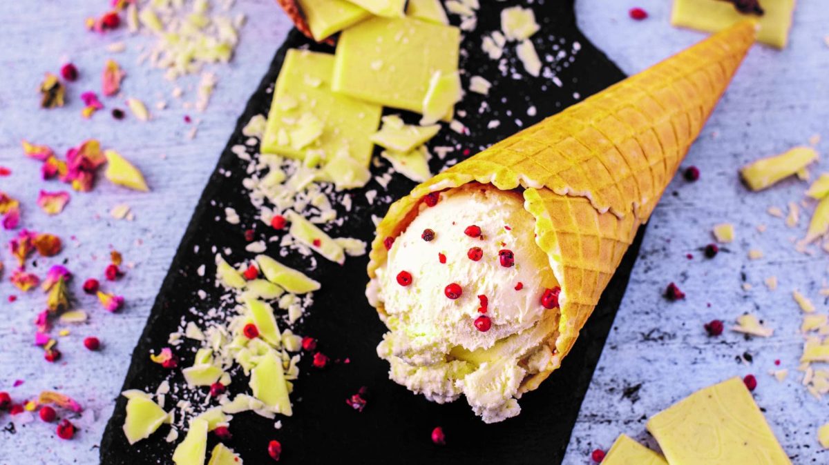 Eine Eiswaffel mit einer Kugel weißes Schoko-Pfeffer-Eis liegt auf einer Schieferplatte, drumherum zerbrochene weiße Schokolade und rote Pfefferkörner.