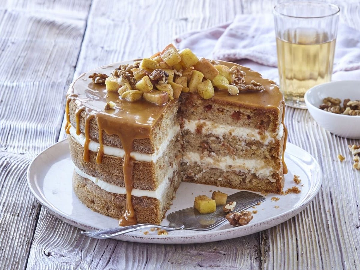 Auf einem Teller steht eine Apfel-Karamell-Schichttorte. Sie ist mit Äpfeln, Zimt und Walnüssen bedeckt.