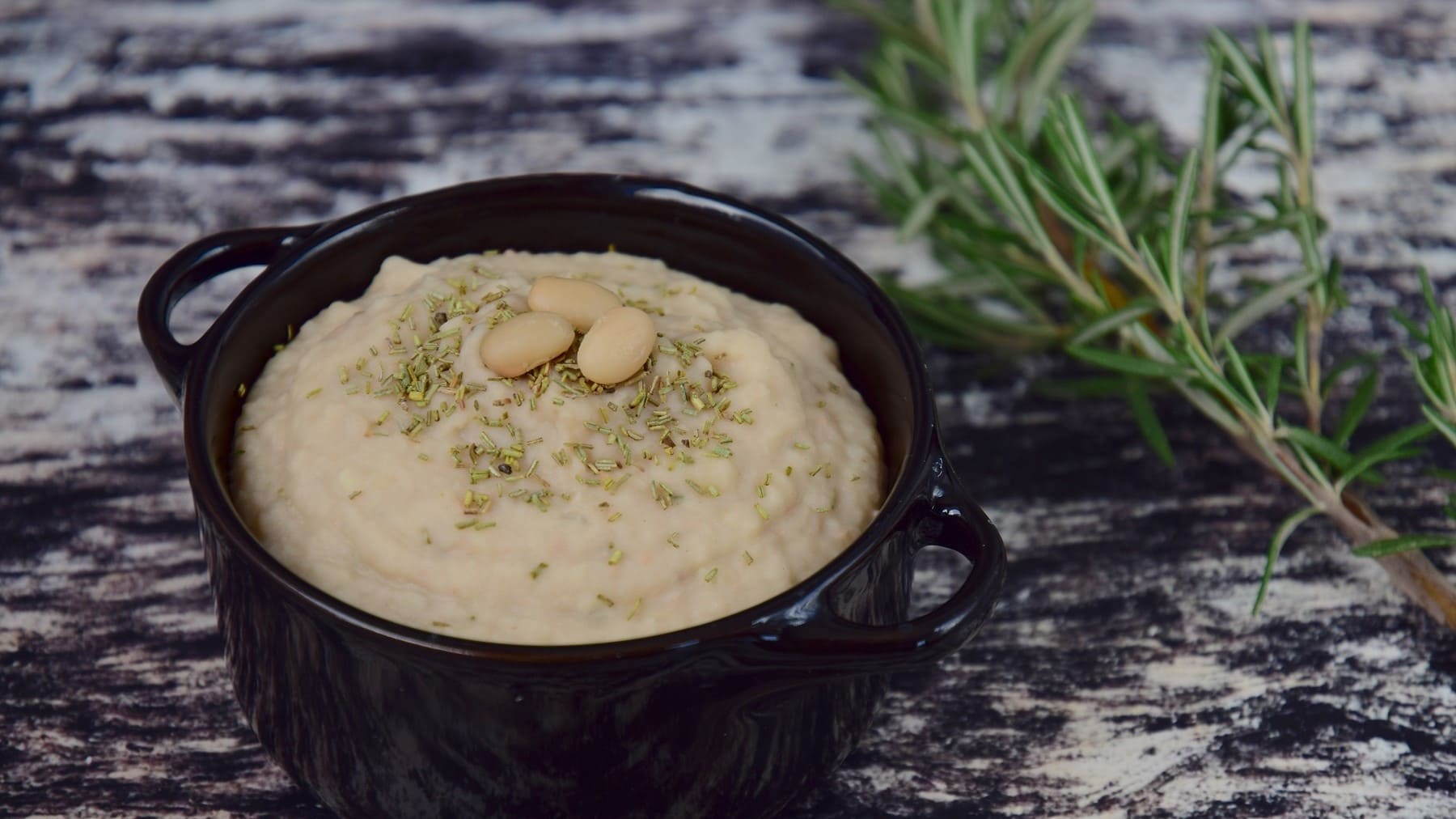 Cremige Bohnen-Paste mit Knoblauch, garniert mit Rosmarin und serviert in einer kleinen schwarzen Schale auf einem Marmortisch.