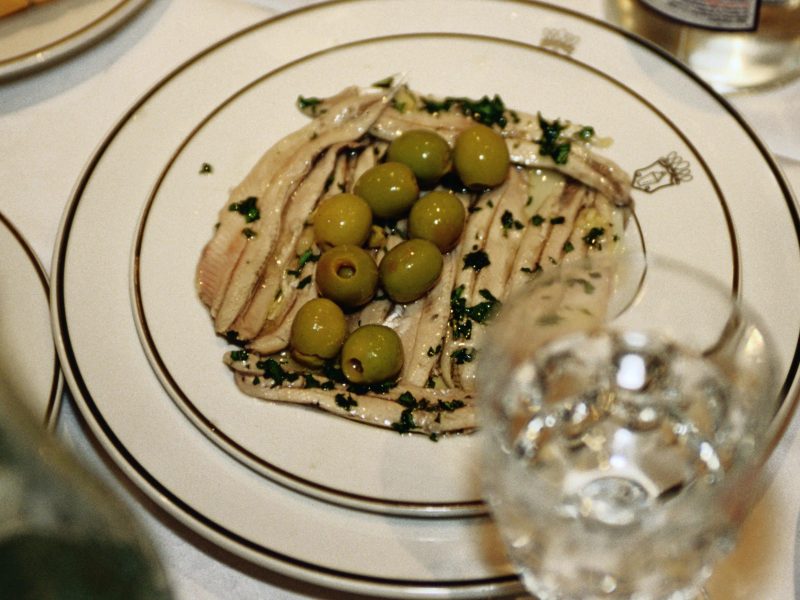 Boquerones en Vinagre eingelegte Sardellen auf weißem Teller und weißem Tisch. Daneben ein Glas Wasser und weitere Tapas am Bildrand. Draufsicht.