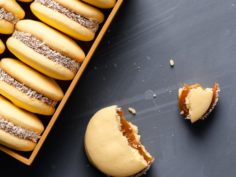 Box mit mehreren Alfajores, daneben ein einzelner in zwei Stücken vor dunklem Hintergrund.