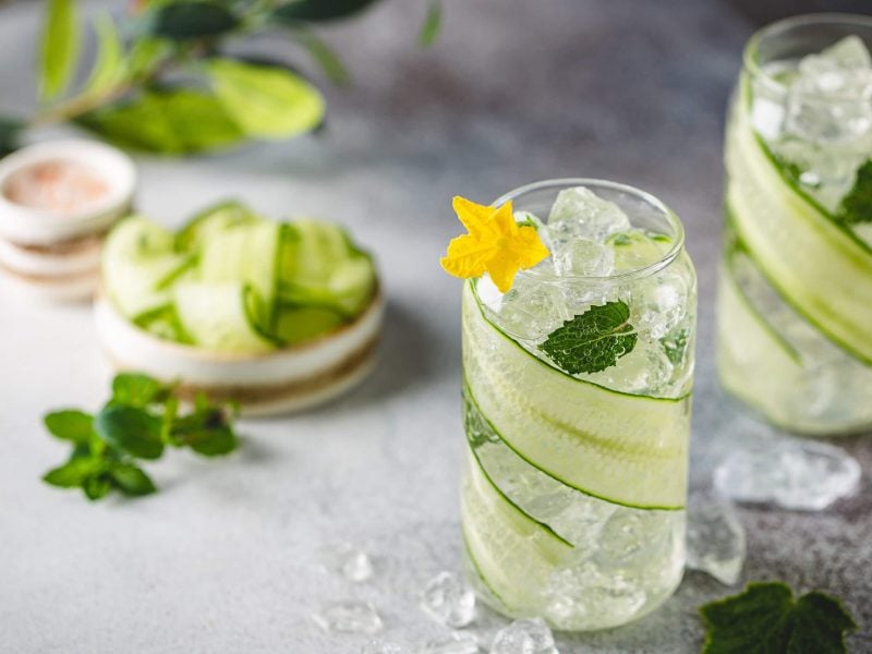 Zwei Gläser mit der leckeren Limonade auf einem grauen Untergrund. Neben den Gläsern liegt Minze und weitere Gurkenstücke.