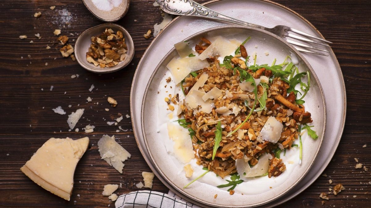 Haferpfanne mit Pfifferlingen und Joghurt, garniert mit Rucola und groben Parmesanstreifen, Walnüssen und Kräutern, serviert auf einem hellen Teller auf dunklem Untergrund.