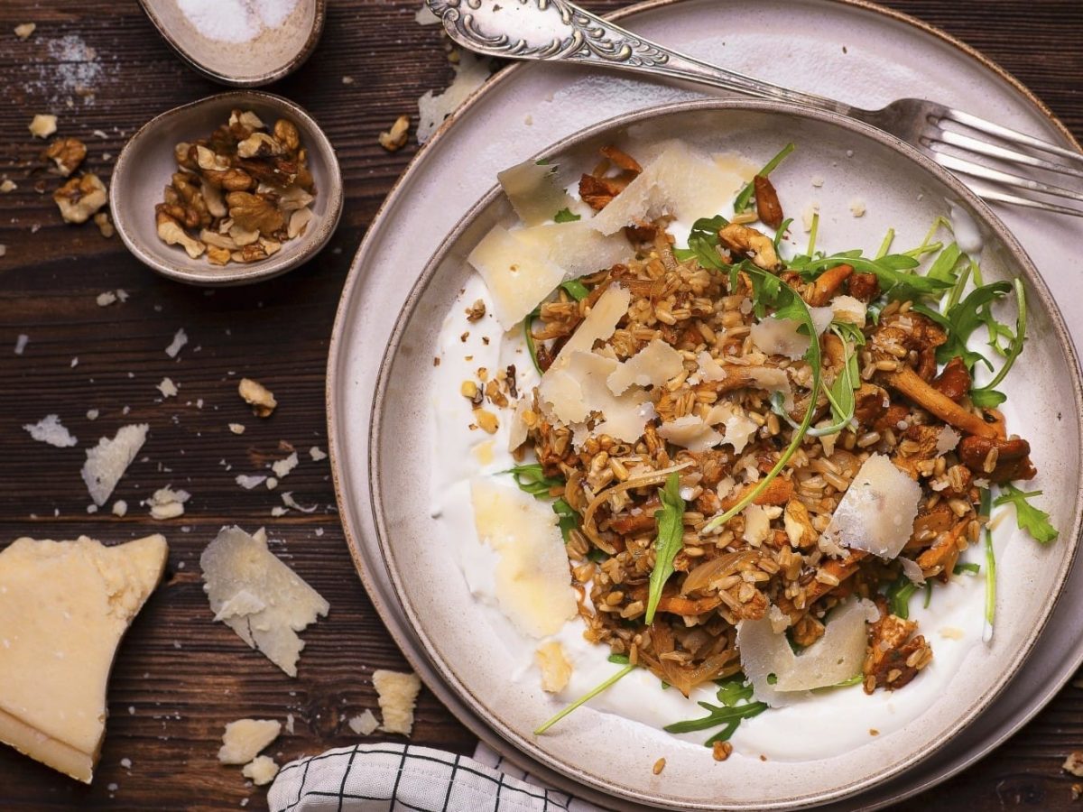 Haferpfanne mit Pfifferlingen und Joghurt, garniert mit Rucola und groben Parmesanstreifen, Walnüssen und Kräutern, serviert auf einem hellen Teller auf dunklem Untergrund.