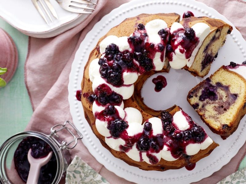 Mit Sahne und Heidelbeerkonfitüre garnierter Heidelbeer-Marmorkuchen, serviert auf einem weißen Teller auf rosanem Tischtuch mit zusätzlicher Konfitüre und einem Strauß Tulpen.