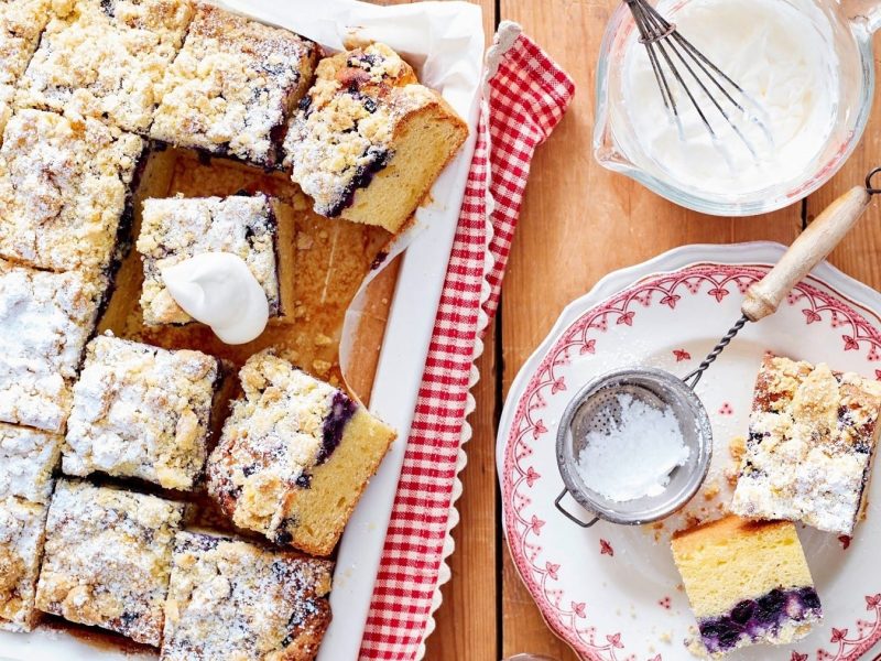 Heidelbeerkuchen mit Orangenstreuseln, als Blechkuchen in kleine Stücke angeschnitten auf einer Form serviert, mit Puderzucker bestreut und mit geschlagener Sahne gereicht.