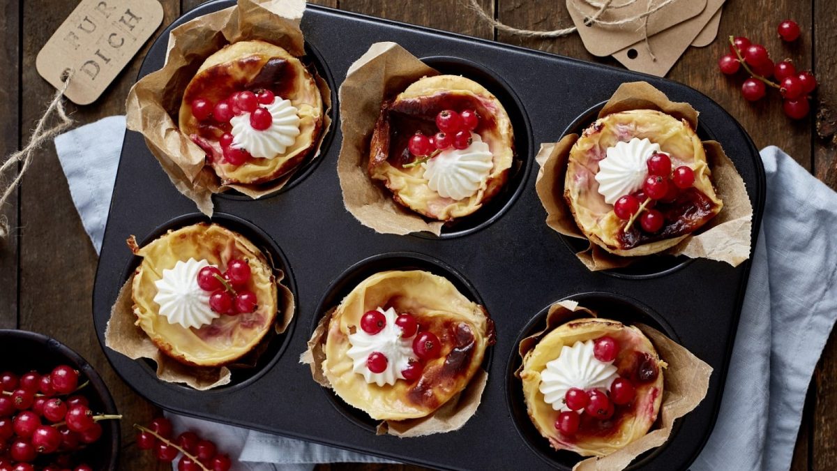 In einer Muffinform sind sechs Johannisbeer-Mandel-Törtchen zubereitet. Sie sind mit Schlagsahne verziert.