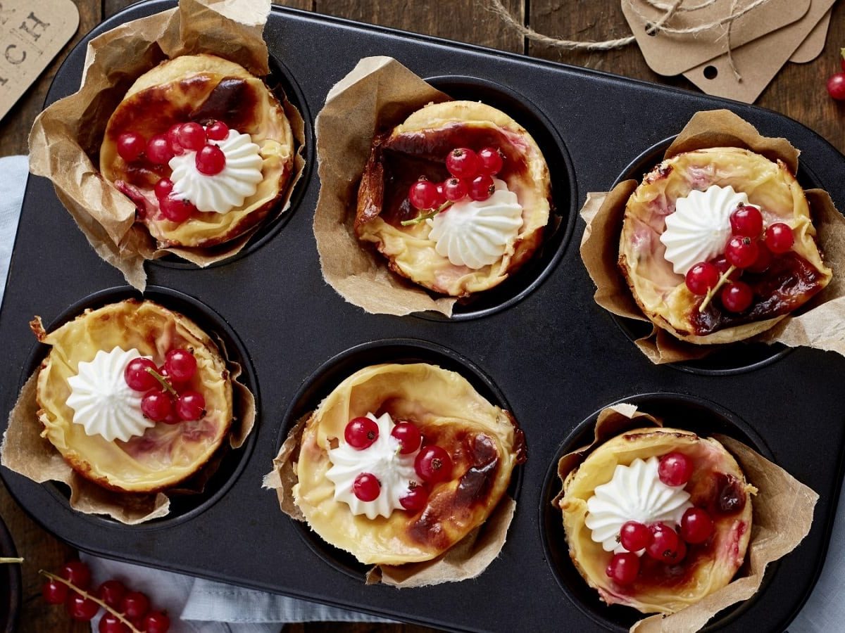 In einer Muffinform sind sechs Johannisbeer-Mandel-Törtchen zubereitet. Sie sind mit Schlagsahne verziert.