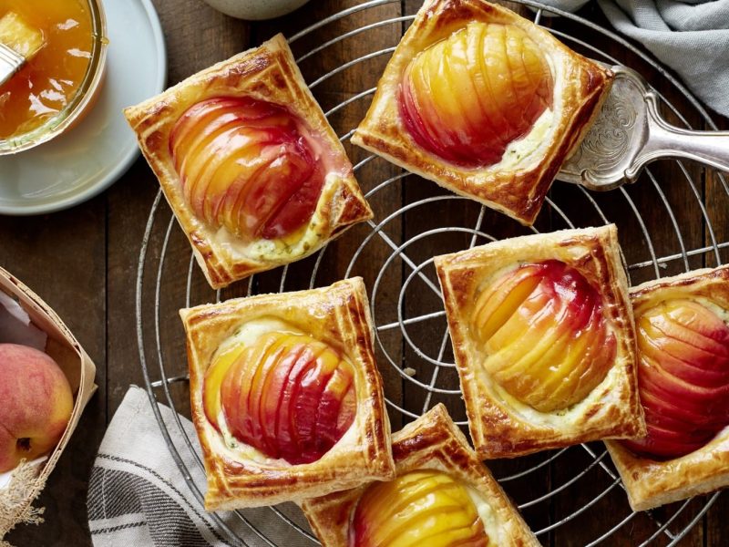 Knusprige Pfirsich-Teilchen, bestehend aus Blätterteig-Taschen mit aufgefächerten halben Pfirsichen, die mit Pfirsichkonfitüre bestrichen und auf einem Kuchenrost auf einem Holztisch angerichtet sind.